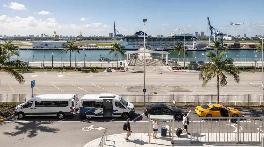 Cruise terminal pickup curb with shuttle vans, a black sedan and a yellow taxi, a cruise ship docked behind and a plane climbing overhead